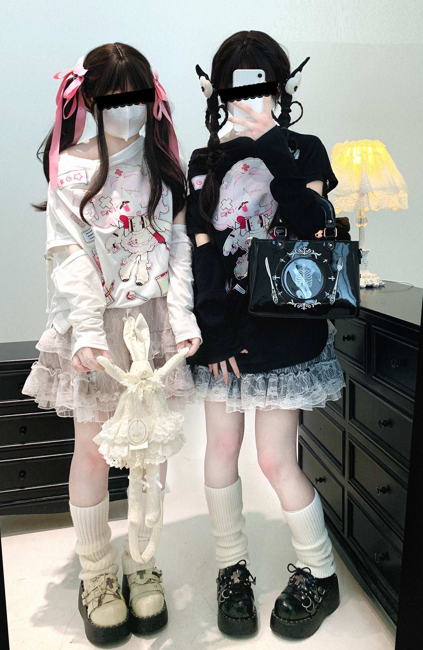 Two individuals in gothic Lolita fashion standing in a room with a lamp in the background.