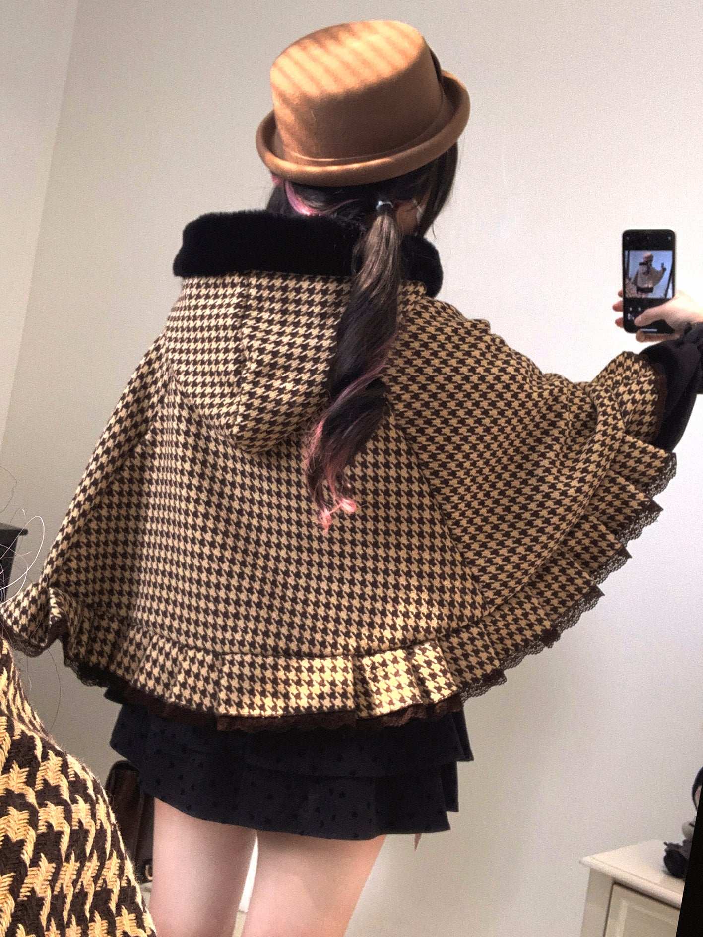 Person wearing a houndstooth patterned cape and brown hat, taking a mirror selfie.