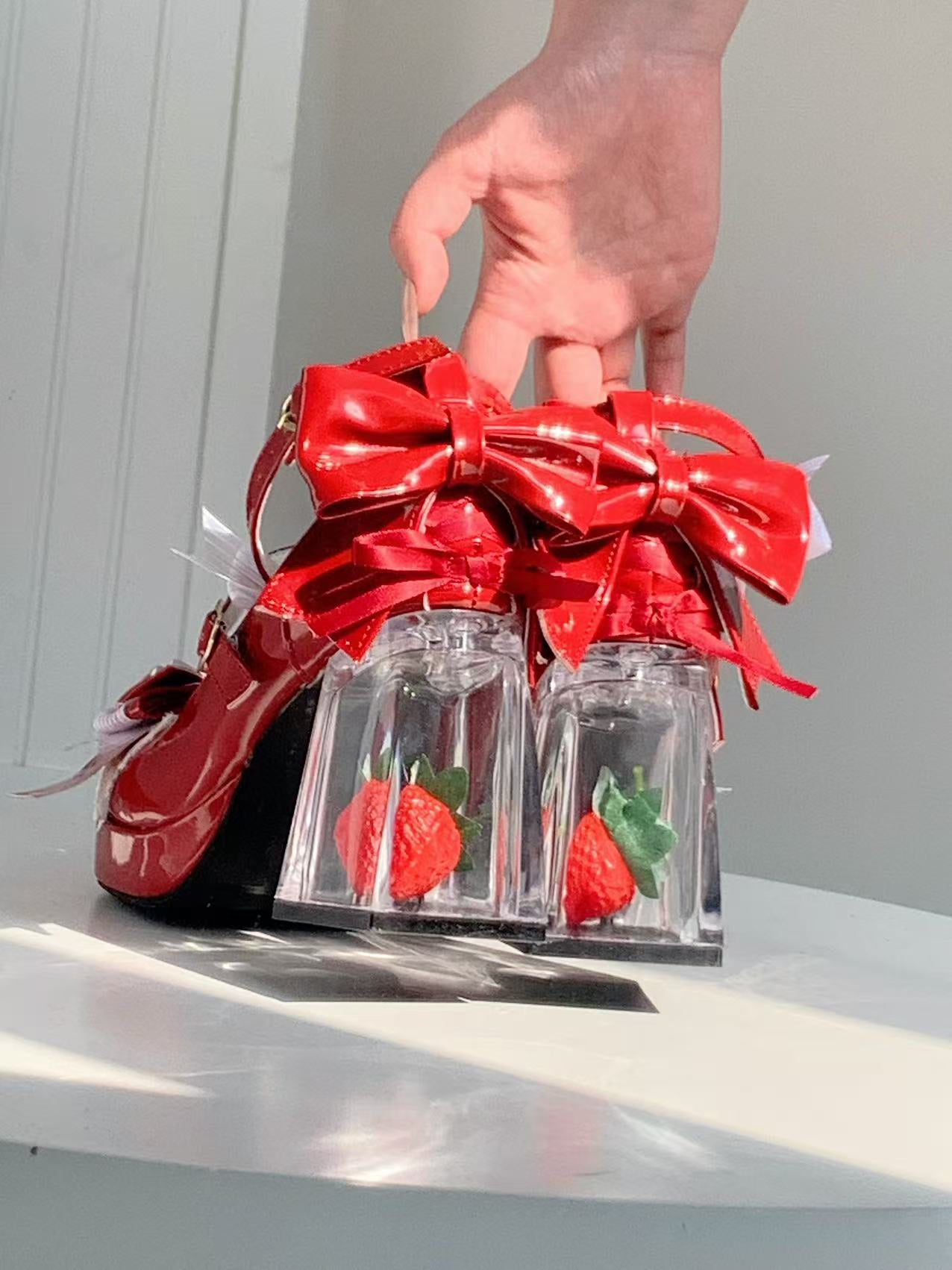 Red shoes with a large bow and clear heels holding strawberries on a white surface.