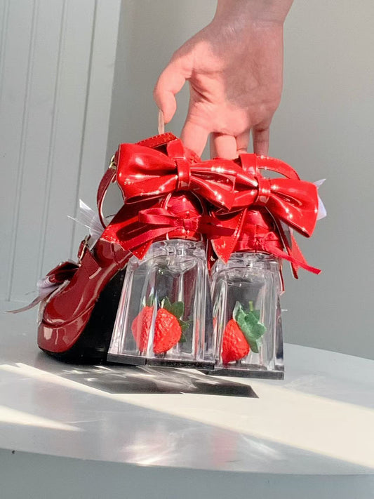Red shoes with a large bow and clear heels holding strawberries on a white surface.