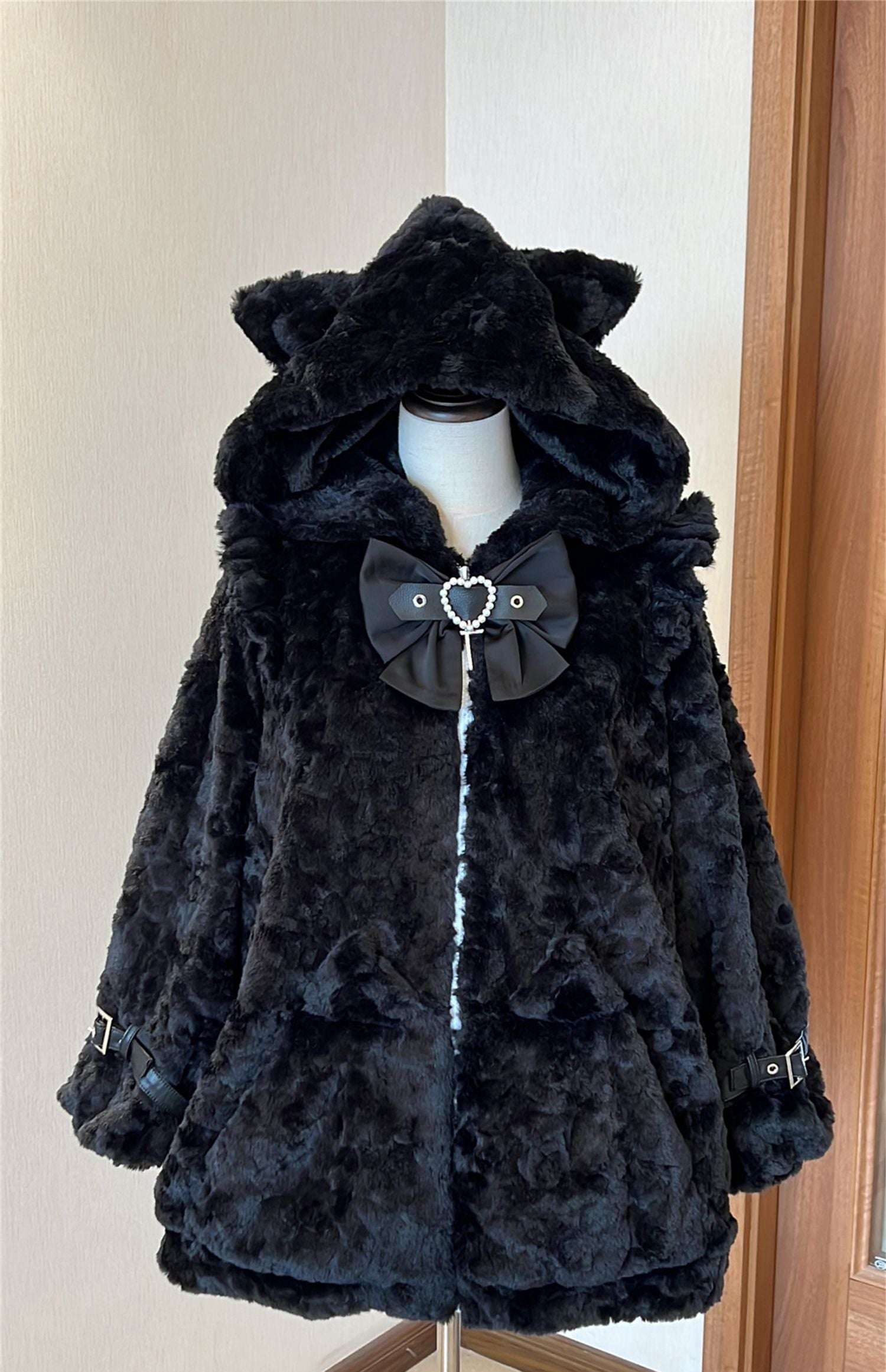 Black faux fur coat with a large bow on a mannequin against a neutral wall.