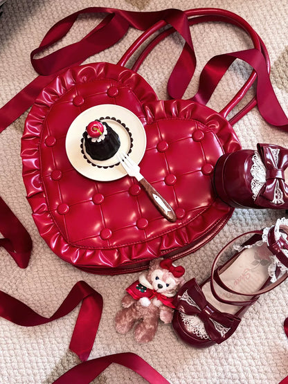 Red heart-shaped bag with a cake design, teddy bear, and shoes on a textured surface.