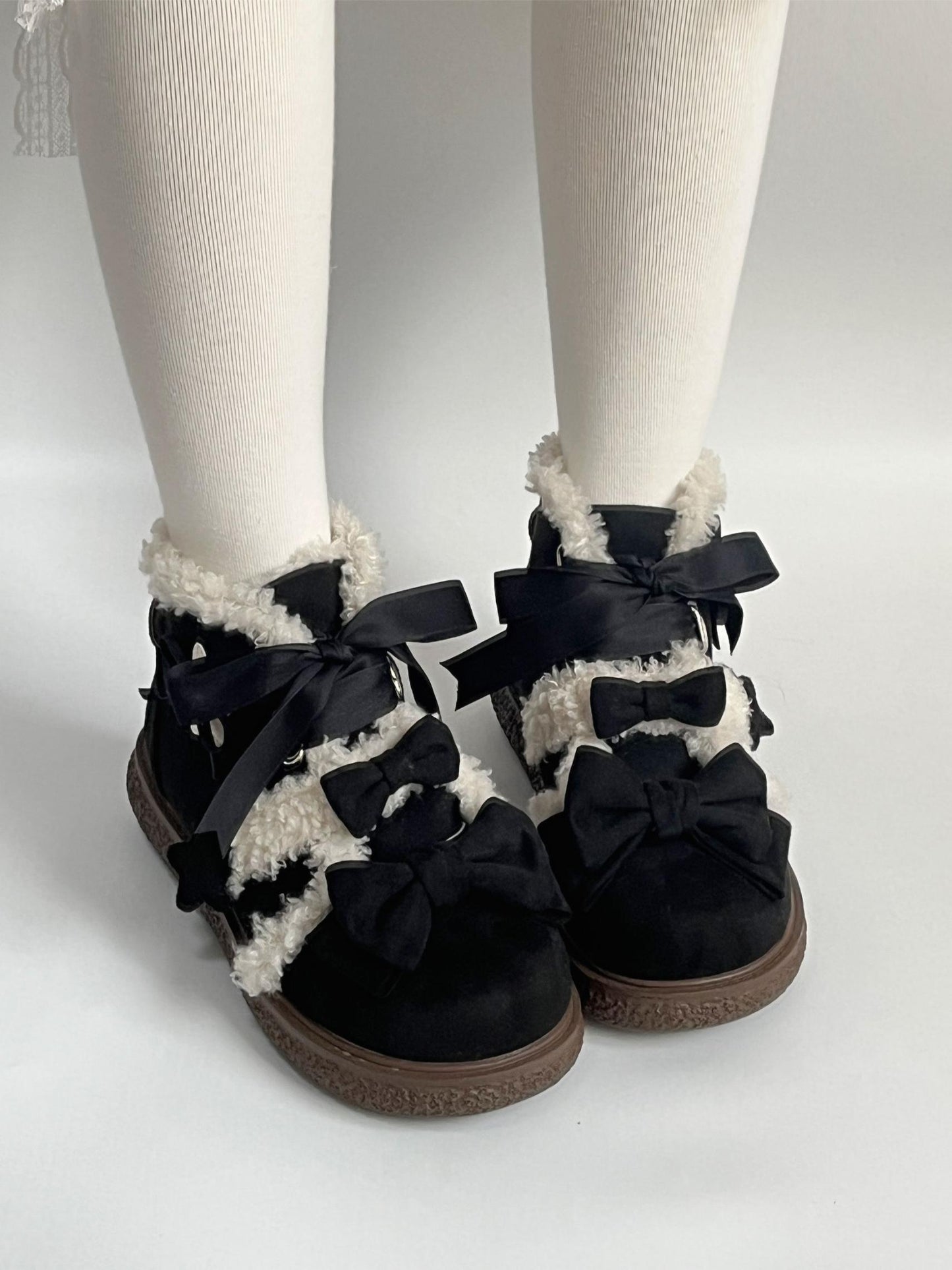 Black lolita style boots with bows worn with white tights on a light gray background