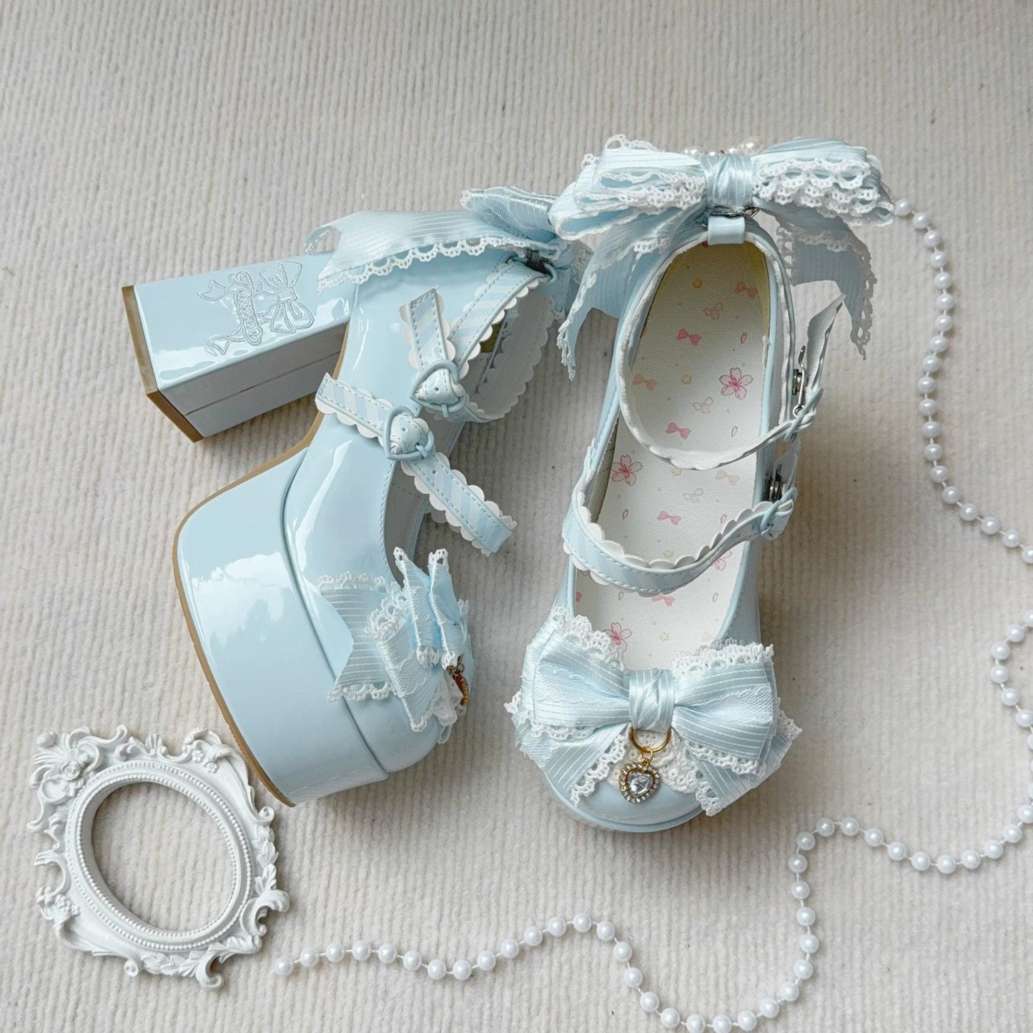 Pair of light blue high-heeled shoes with decorative bows and lace on a beige surface.