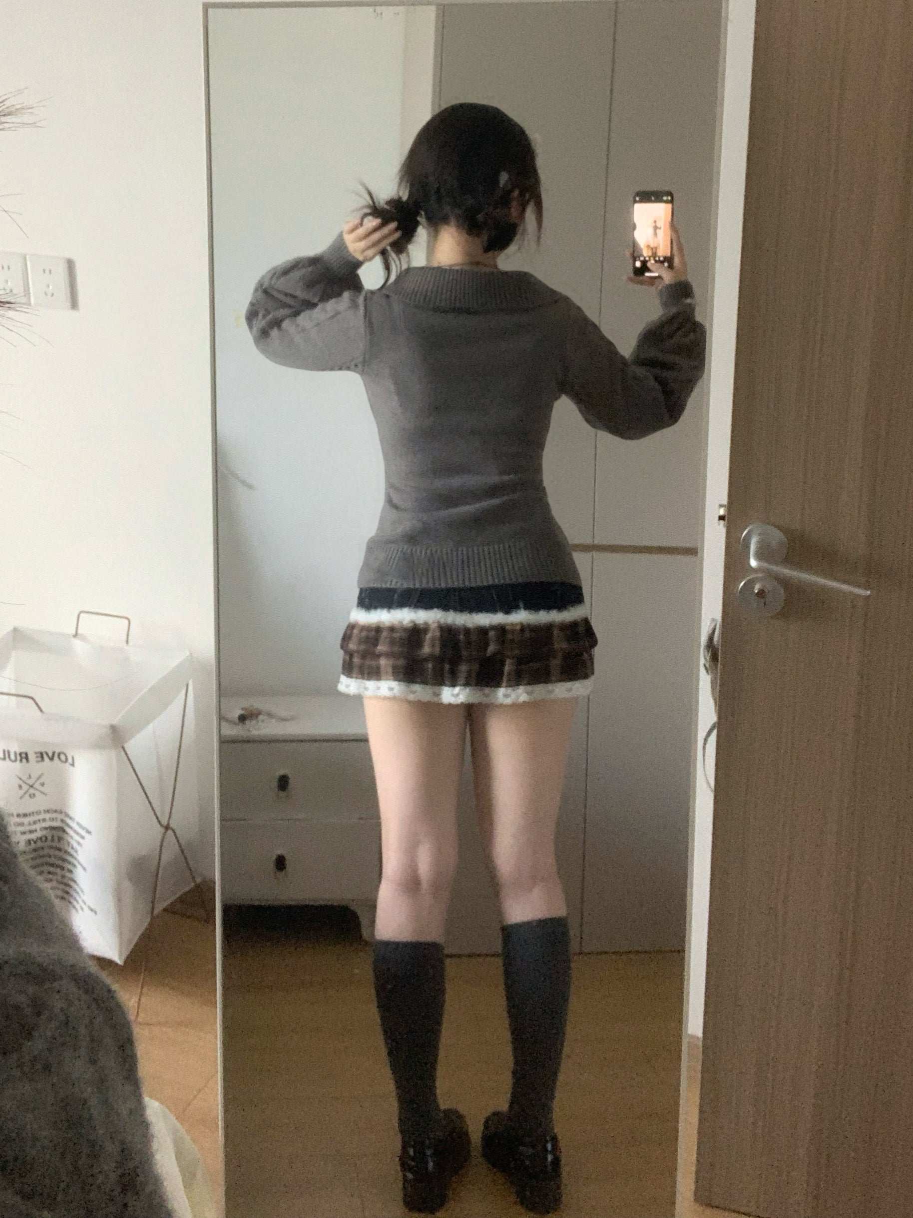Person taking a mirror selfie wearing a gray sweater, patterned skirt, and black boots.