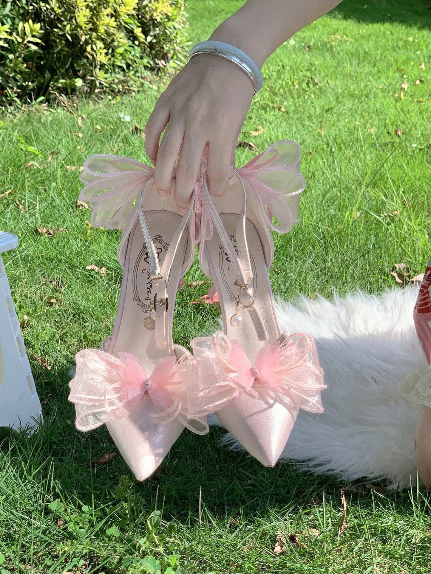 White high-heeled shoes with pink ribbons held by a person on grass