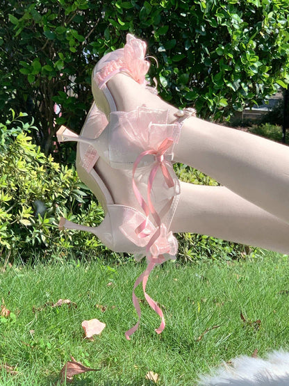 Pink high-heeled shoes with pink ribbons and bows worn outdoors on a grassy area with greenery.