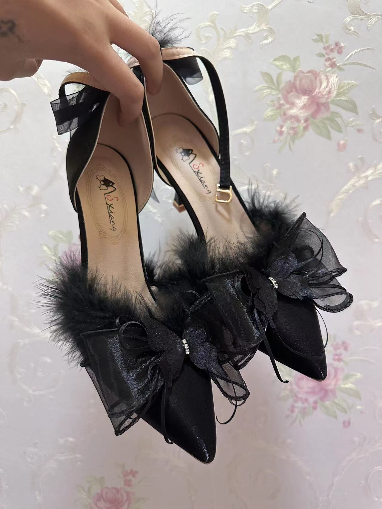 Black high-heeled shoes with feather details held against a floral wallpaper background