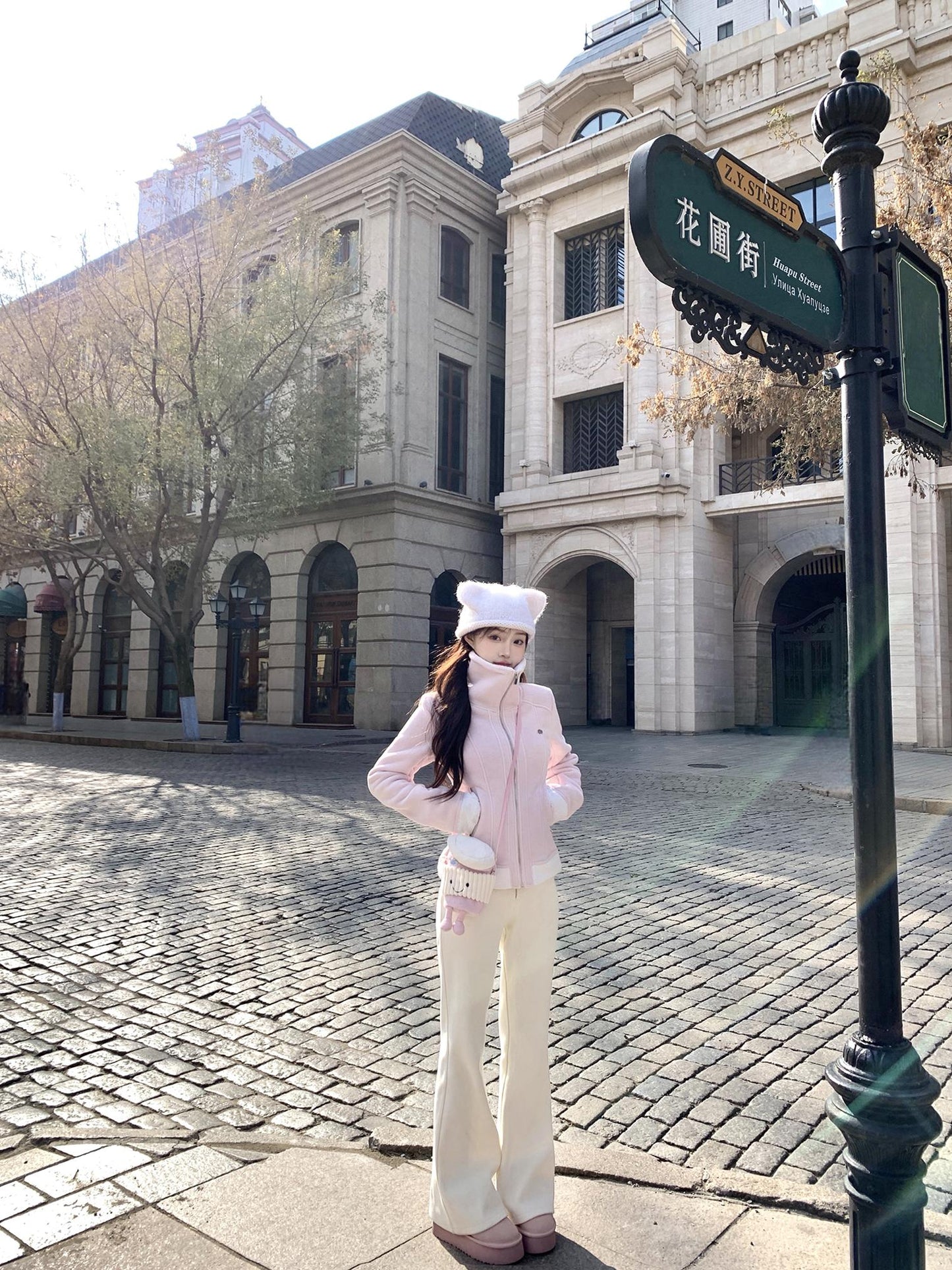 Woman in a pink coat and white hat standing on a cobbled street with classical architecture in the background.