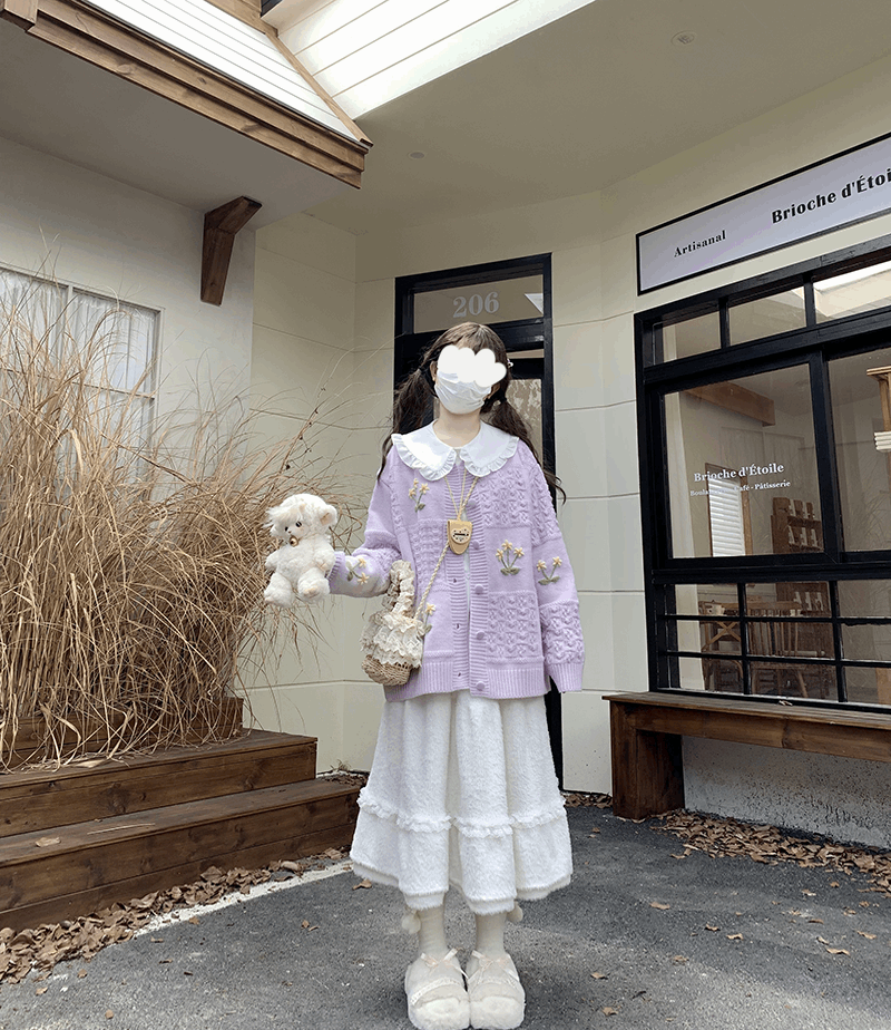 Person in a light purple cardigan and white skirt holding a teddy bear outside a building.