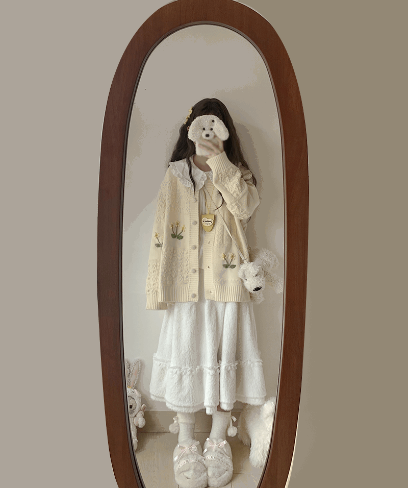 Person wearing a cream-colored cardigan with floral patterns in front of an oval mirror.