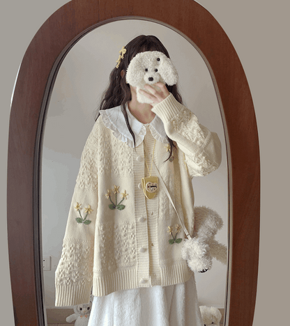 Person wearing a cream-colored cardigan with floral embroidery, holding a small white dog toy.