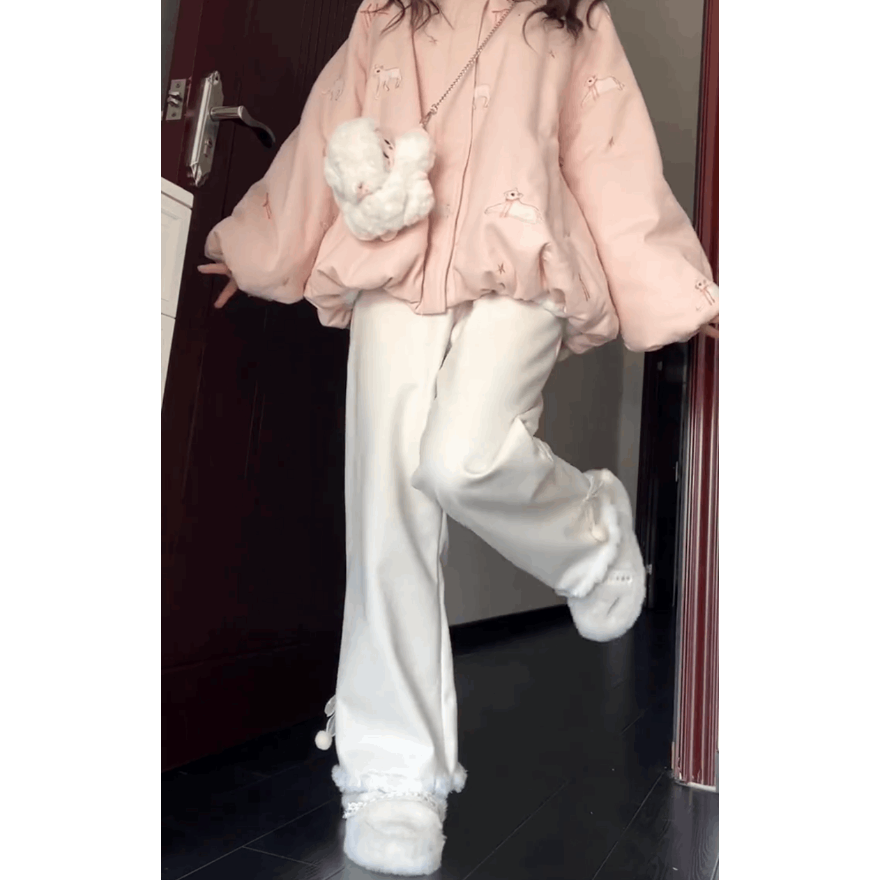 Person wearing a pink outfit with white pants and shoes, holding a white plush toy, standing in a doorway.