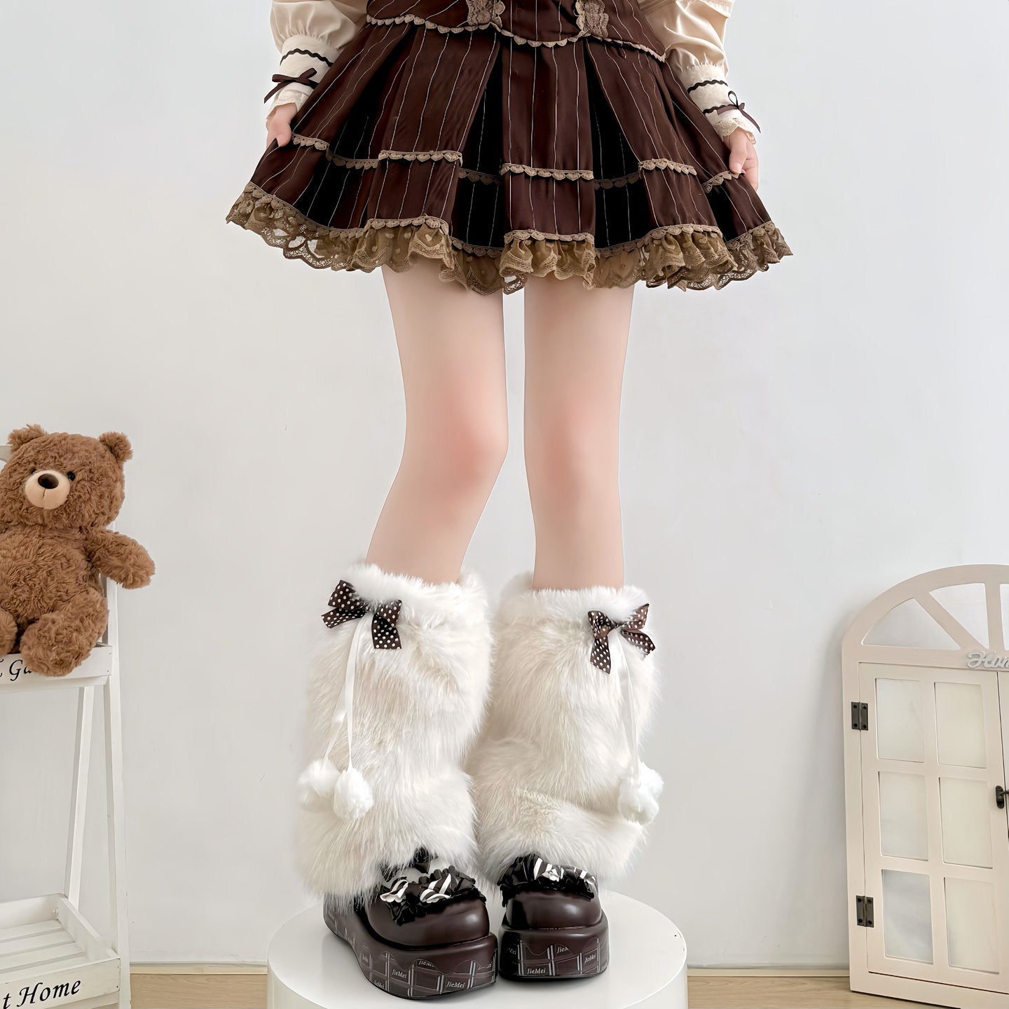 White furry leg warmers with bows worn over dark shoes, paired with a brown skirt and teddy bear in the background.