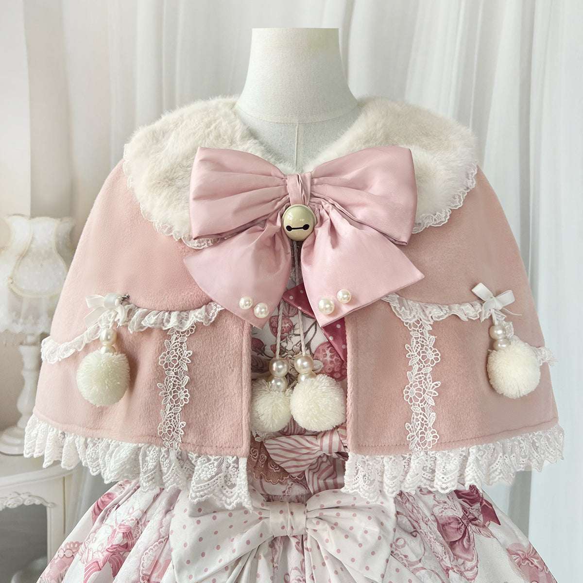 Pink cape with a large bow and decorative elements on a mannequin against a white curtain background.