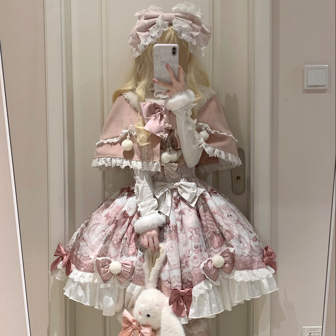 Person wearing a pink and white Lolita-style outfit with a bear plushie, standing in front of a mirror.