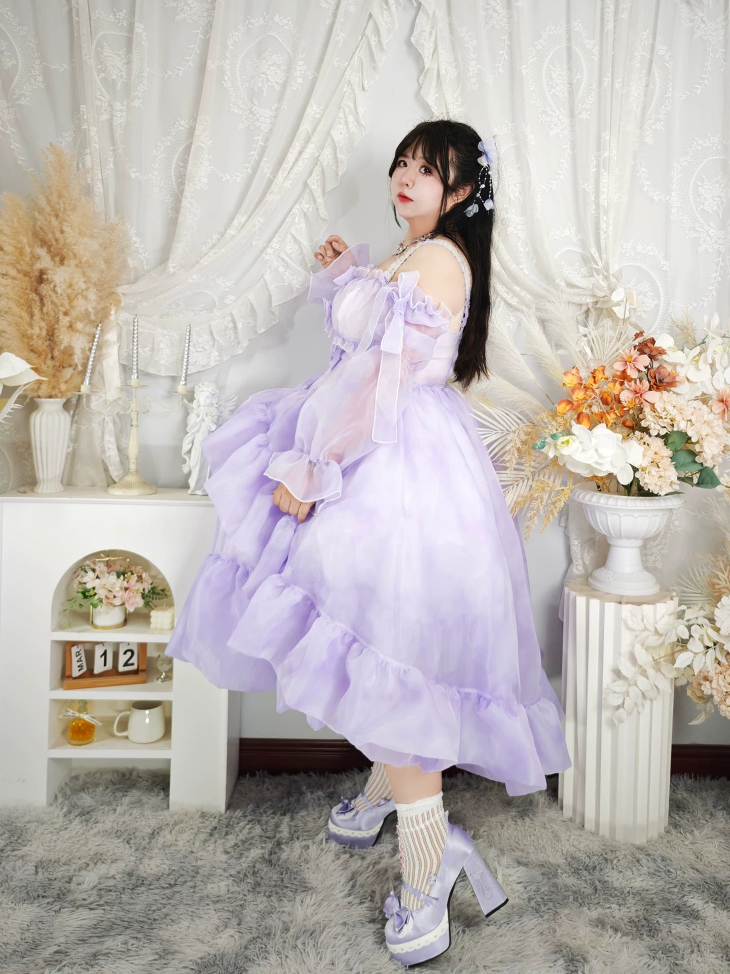 Woman in a lavender dress standing in a decorated room with flowers and decor.