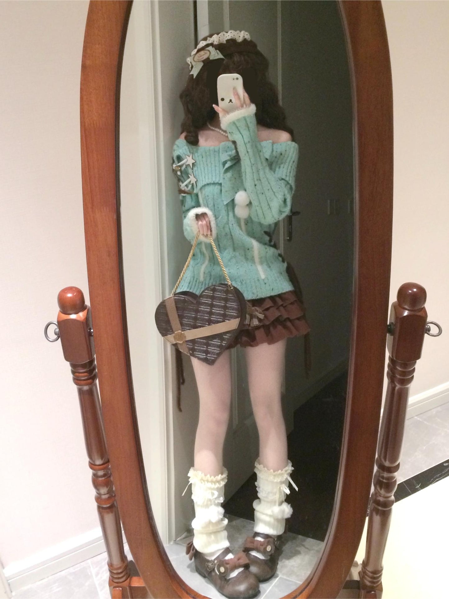 Person wearing a green sweater and skirt, holding a heart-shaped bag, standing in front of a wooden-framed mirror.