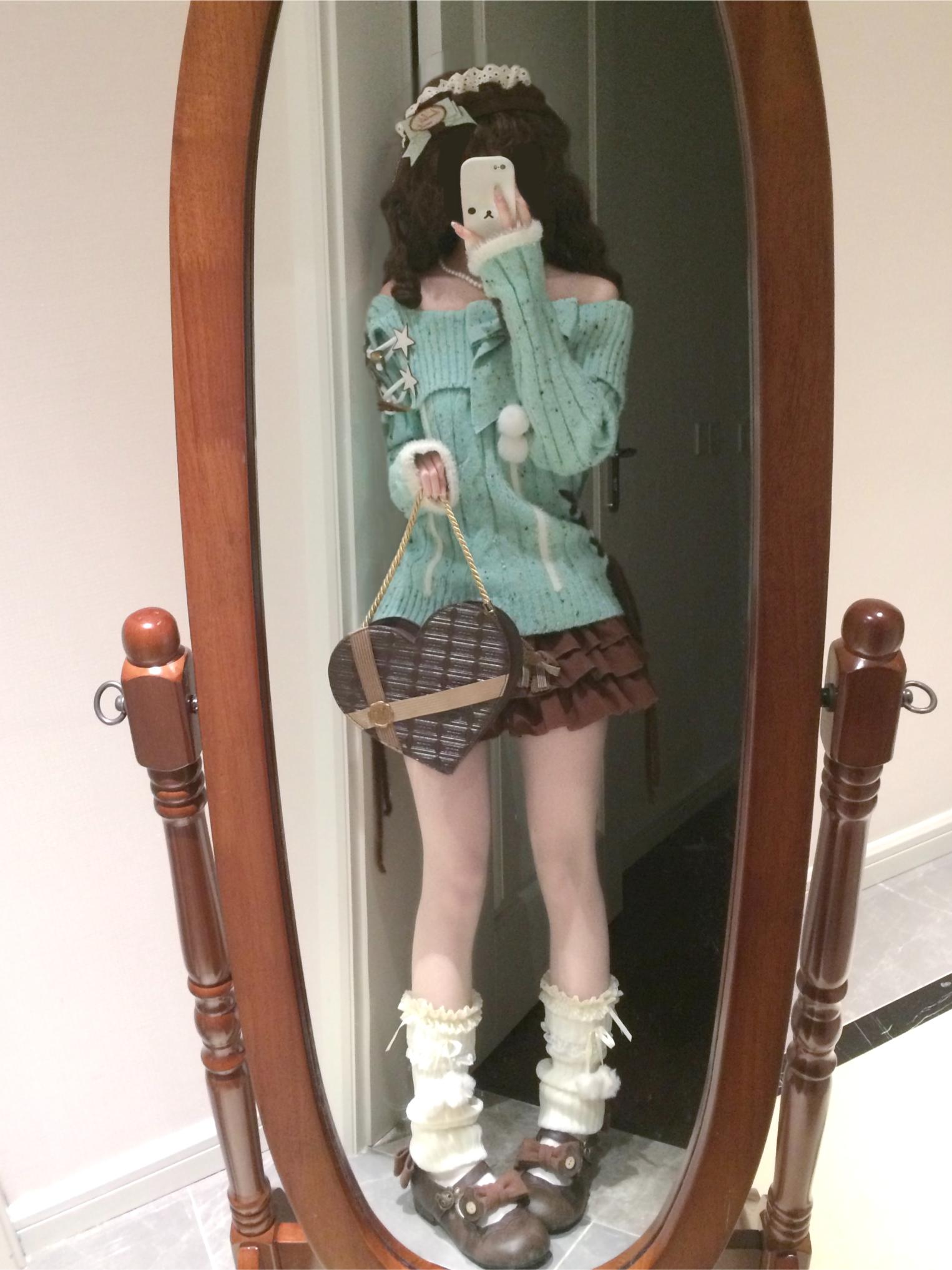 Person wearing a green sweater and skirt, holding a heart-shaped bag, standing in front of a wooden-framed mirror.