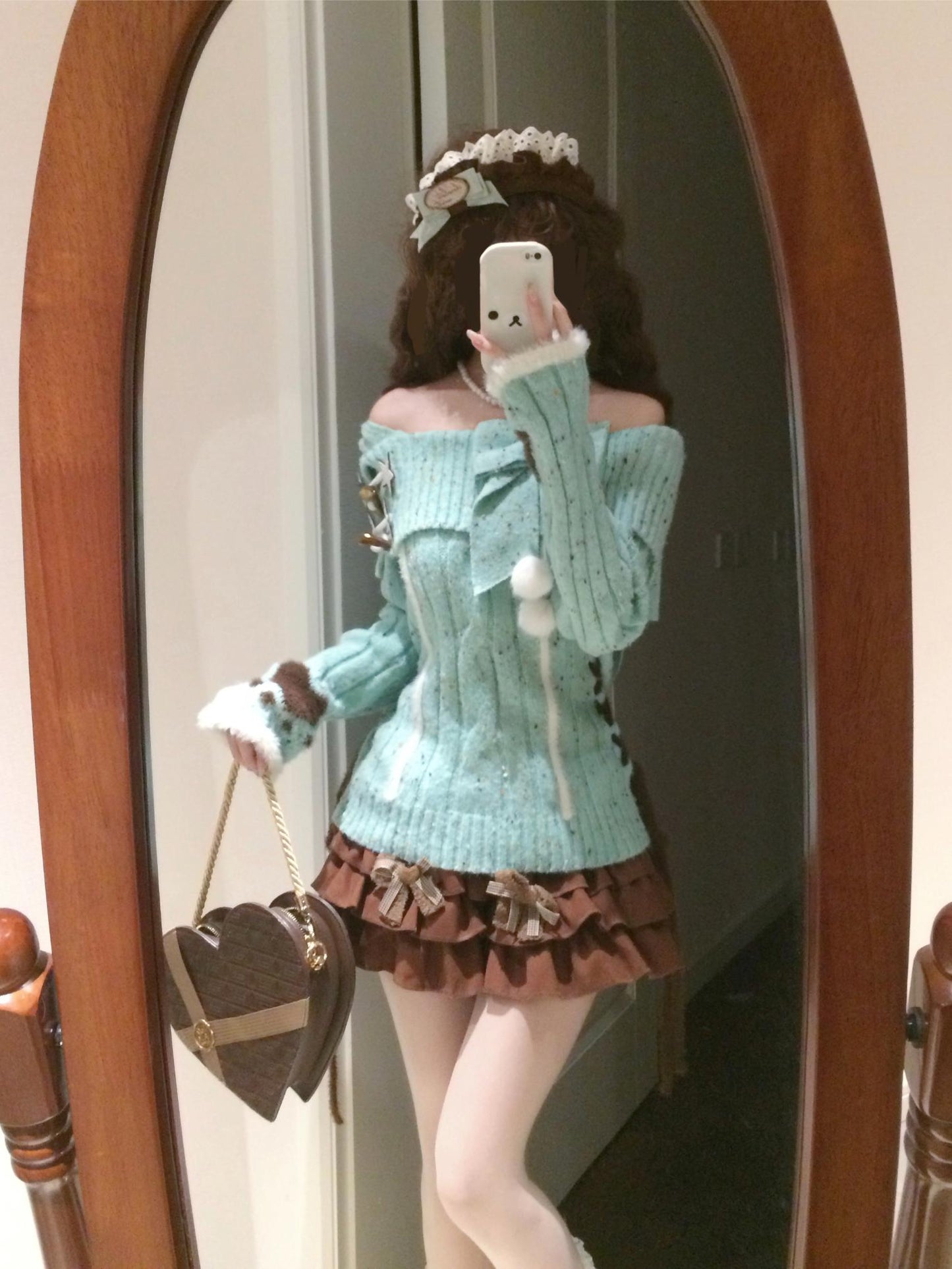 Person wearing a light blue sweater and brown skirt, holding a heart-shaped bag, in front of a mirror.