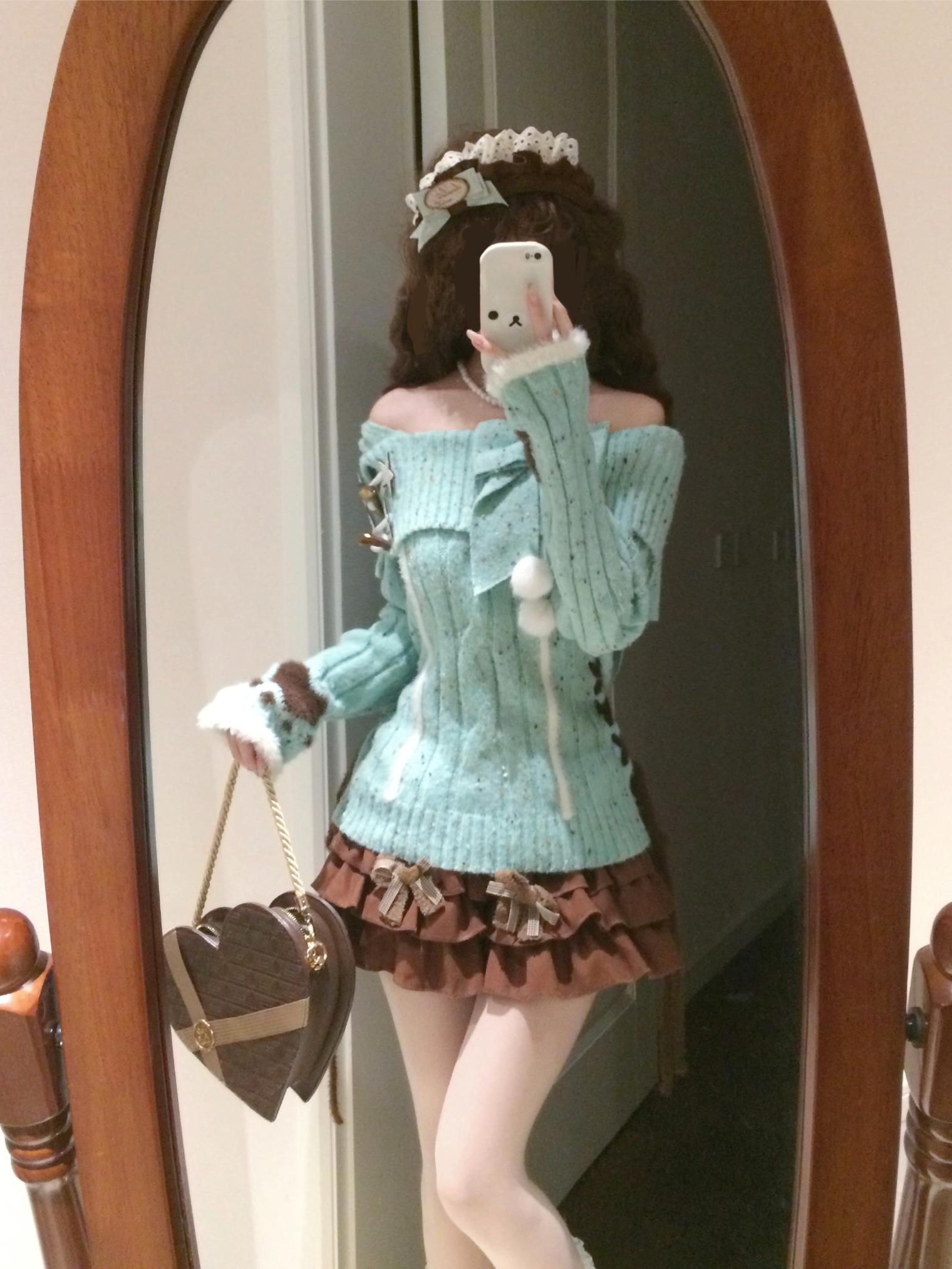 Person wearing a light blue sweater and brown skirt, holding a heart-shaped bag, in front of a mirror.