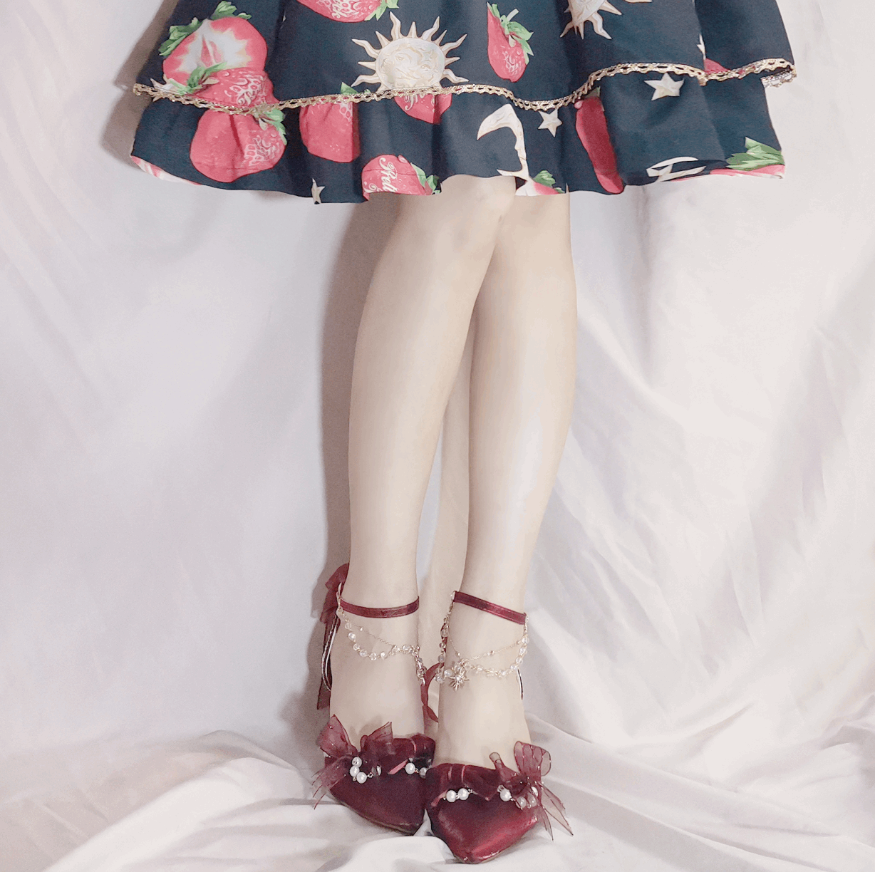 Close-up of a person wearing a floral dress, white stockings, and red shoes with decorative bows on a light background.