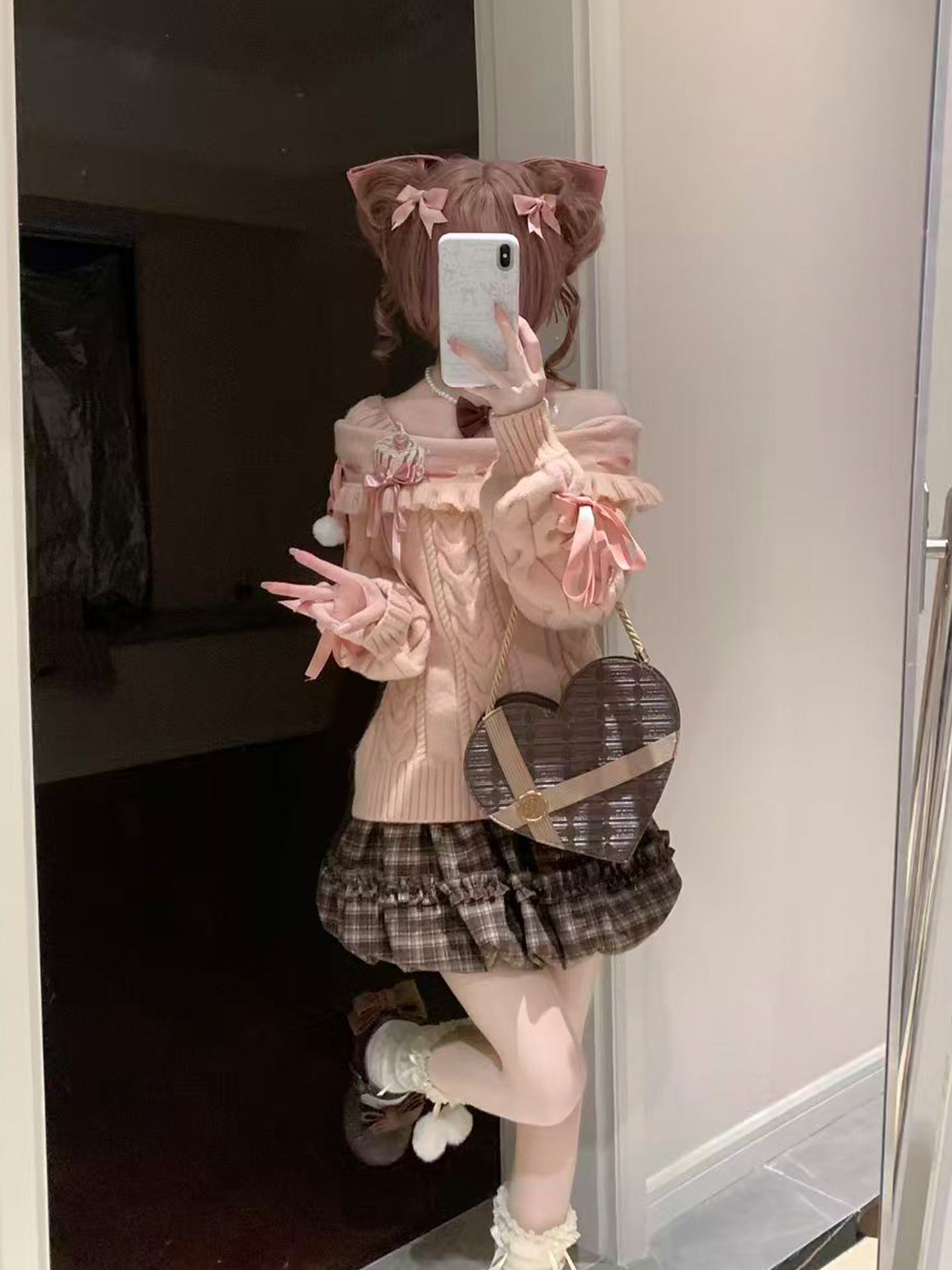 Person wearing a pink sweater and plaid skirt with a heart-shaped bag, taking a mirror selfie.