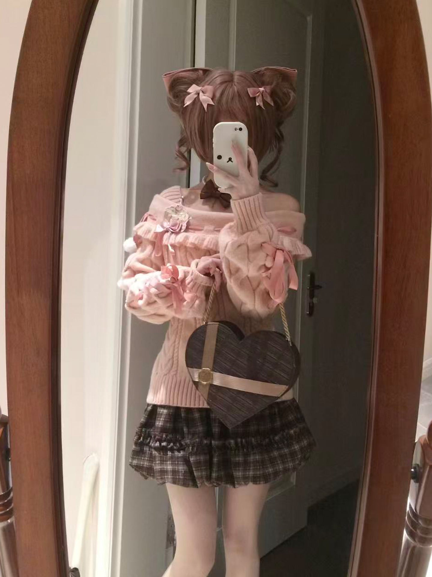 Person wearing a pink sweater and plaid skirt with a heart-shaped bag, standing in front of a mirror.